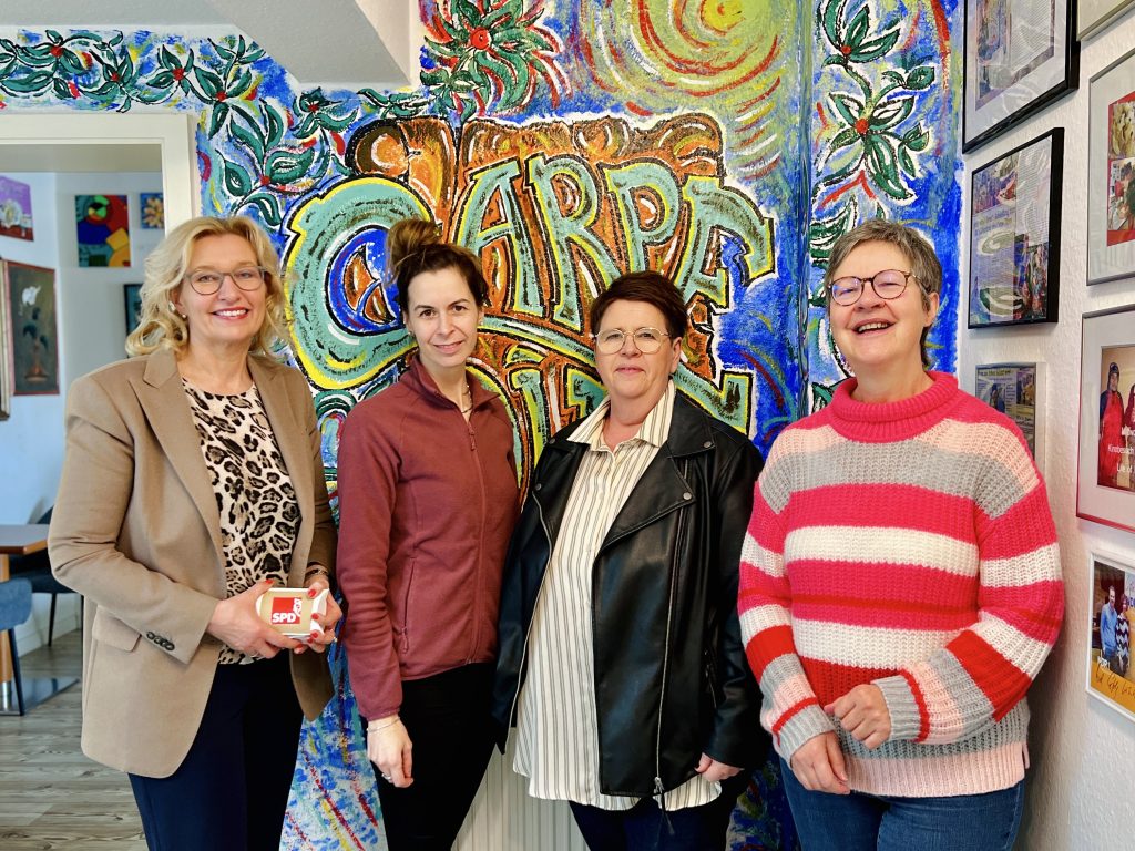Vier Frauen stehen lächelnd in einem Raum mit farbenfroher Kunst an den Wänden und freuen sich über gesammelte Spenden.
