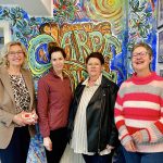 Vier Frauen stehen lächelnd in einem Raum mit farbenfroher Kunst an den Wänden und freuen sich über gesammelte Spenden.
